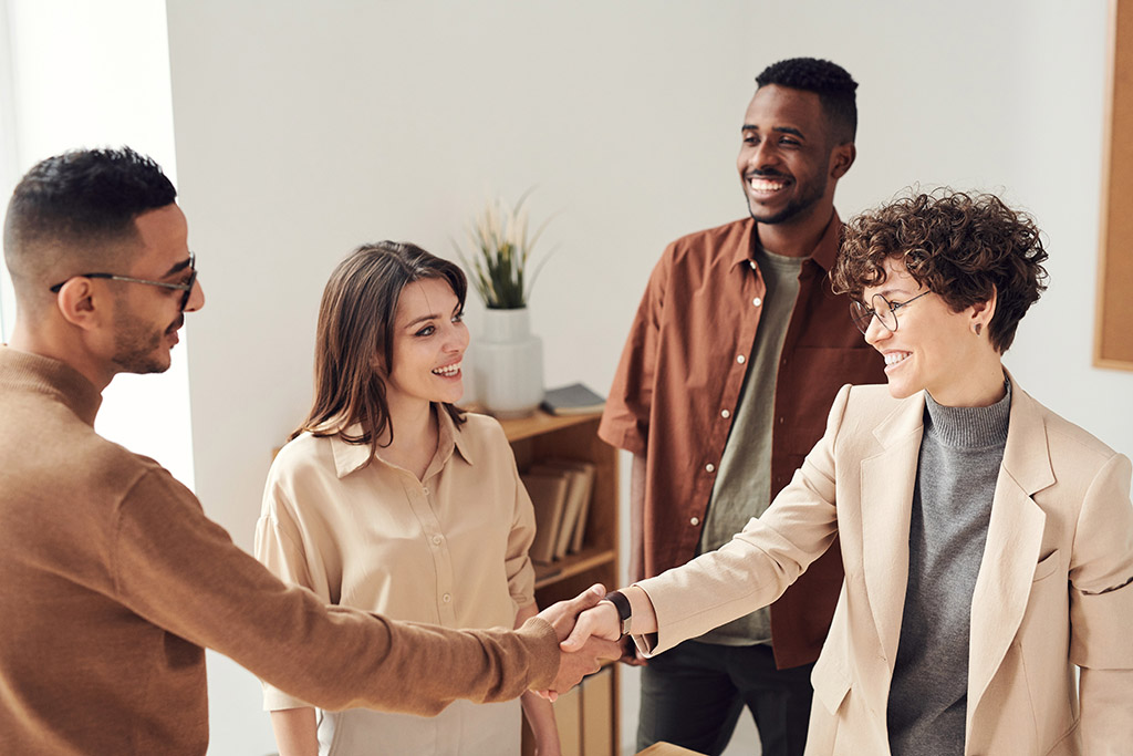 Four people of different colors standing while smiling and two of them are doing handshakes.